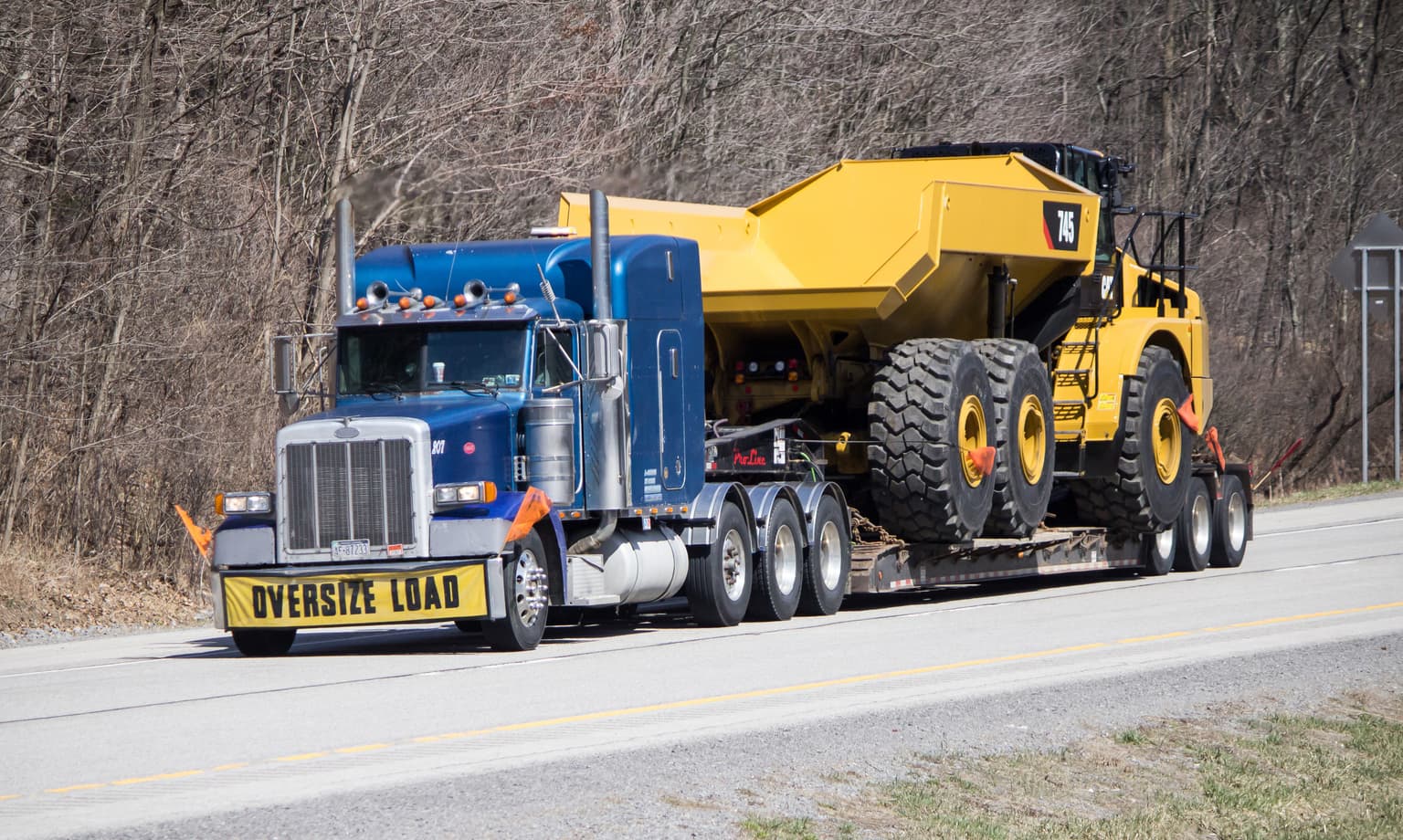 Image related to blog title Blue and yellow semi-truck hauling a large piece of construction equipment on a highway.