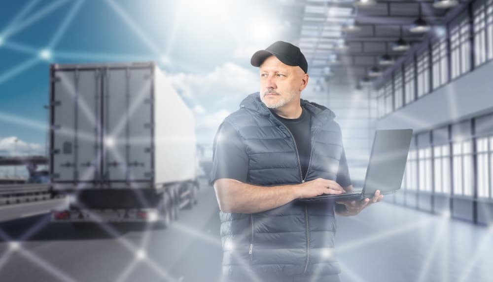 Image related to blog title Trucking company dispatcher using a laptop to coordinate logistics with a truck in the background